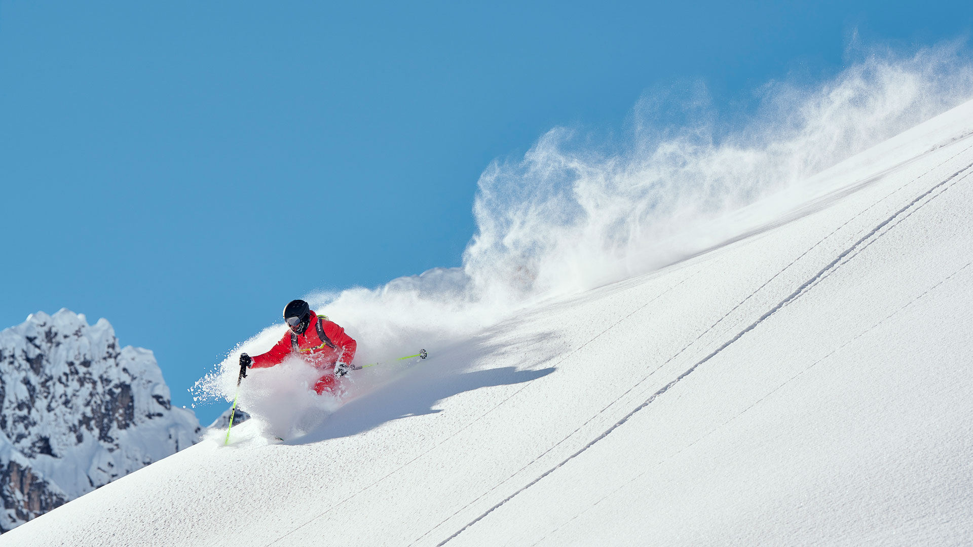 Freerider im roten Anzug zieht tiefe Powderkurve; blauer Himmel, Gipfel im Hintergrund.